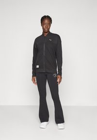 Sort athletic jakke med lynlås, lange ærmer og forlommer. Kombineret med sorte tætsiddende bukser med logo accenter. Hvide sneakers.