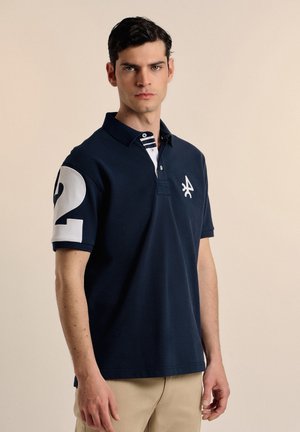Camiseta polo azul marino de manga corta, con acentos blancos y el número "2" en la manga izquierda. Presenta un collar a rayas y un logotipo bordado.