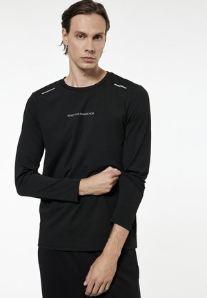 Jeune homme aux cheveux mi-longs portant un t-shirt noir à manches longues avec l'inscription « READY FOR CHANGE NOW », ajustant sa manche sur un fond blanc.