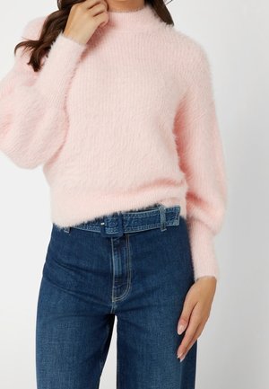 Femme portant un pull doux rose pâle à manches bouffantes associé à un jean bleu foncé taille haute avec une ceinture en jean assortie.