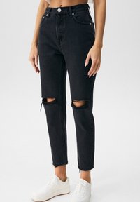 Jeans de mezclilla negra de talle alto, con dos rodillas rasgadas y dobladillos deshilachados, combinados con zapatillas blancas.