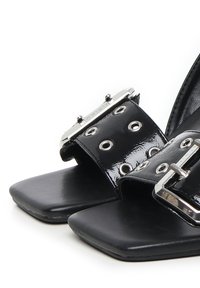 Sandales en cuir verni noir avec une semelle plate, présentant des accessoires en métal argenté, de grands œillets et une large sangle sur le dessus.