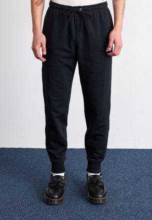 Persona con pantalones jogger negros con cintura de cordón y mocasines negros con borlas, de pie sobre una alfombra azul contra una pared blanca.