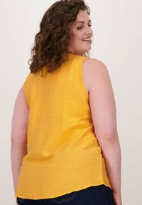 Geel mouwloos topje met een gestructureerde oppervlakte, ronde halslijn en een lichte curve bij de zoom, gecombineerd met donkerblauwe denim jeans.