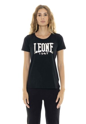 Une femme porte un tee-shirt noir avec l’inscription blanche « LEONE 1947 » sur un fond blanc uni.
