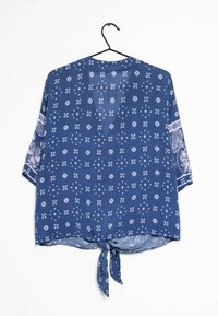 Blouse bleue à manches courtes avec des motifs floraux et géométriques blancs, présentant un détail noué à l'avant, suspendue à un cintre noir sur fond blanc.