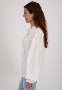 Witte katoenen blouse met een zigzagopen patroon, zachte textuur en gerafelde manchetten; heeft een strikdetail bij de kraag.