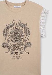 Top beige a maniche corte con frange bianche sulle maniche, design floreale e paisley nero con due uccelli, e testo "VAI CON IL FLUSSO" sul davanti.