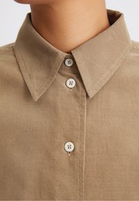Beige Cordhemd mit klassischem Kragen, weißen Knöpfen und feiner vertikaler Rippstruktur. Fokus auf den Details von Kragen und Knopfleiste.