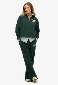 Felpa verde in pile con zip a quarto, scritta bianca e vestibilità comoda; pantaloni abbinati con logo. Maglia a righe indossata sotto.