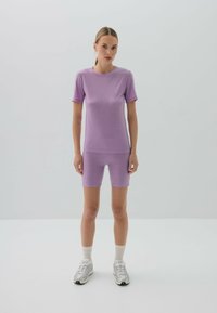 Fioletowy dopasowany top i pasujące szorty rowerowe wykonane z gładkiego, elastycznego materiału. Noszone z białymi trampkami i skarpetkami w kolorze kremowym sięgającymi do kolan.