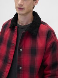 Κόκκινο και μαύρο πουκάμισο-ζακέτα με καρό σχέδιο και μαύρο φλις γιακά, με κουμπιά τύπου σναπ και απαλή υφή. Φορεμένο πάνω από σκούρο γκρι πουκάμισο.