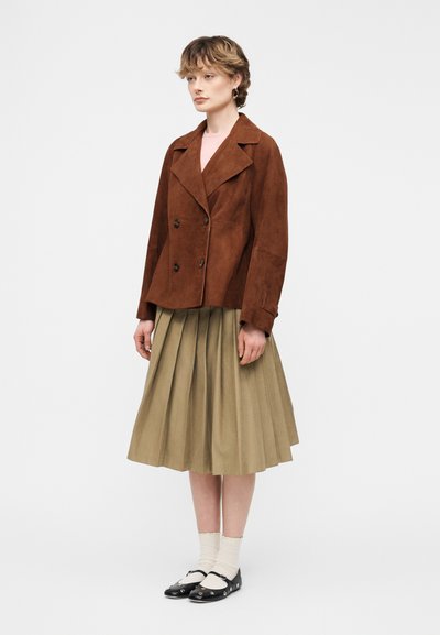 Νεαρή γυναίκα που φοράει καφέ σακάκι με διπλή σειρά κουμπιών, μπεζ πλισέ φούστα, λευκές κάλτσες και μαύρα επίπεδα παπούτσια, στέκεται μπροστά σε μια απλή φόντο.