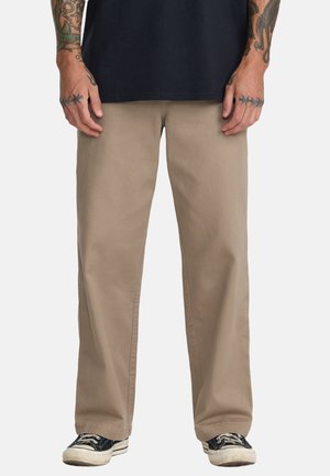 Homme portant un pantalon beige à jambes droites, des baskets noires à semelles blanches et une chemise noire, debout avec les bras tatoués visibles.