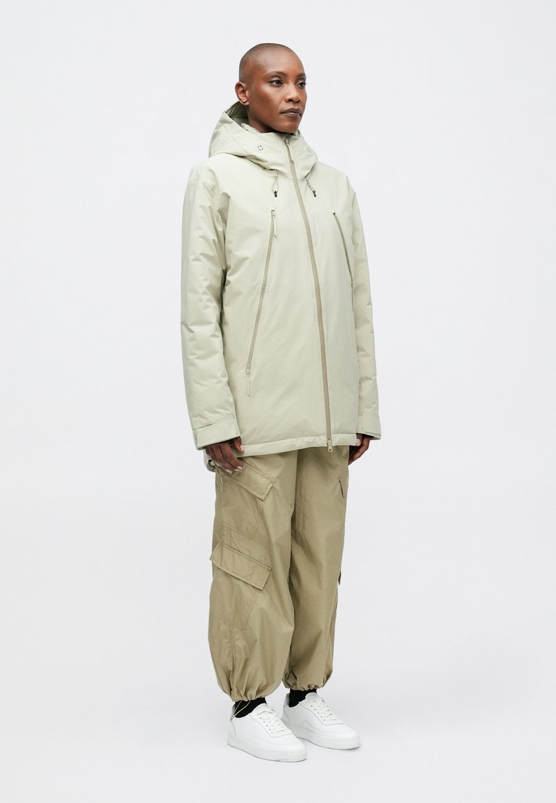 Beige isolierte Jacke mit Kapuze, Reißverschlusstaschen und einer leicht lockeren Passform, kombiniert mit khakifarbenen Cargo-Hosen und weißen Turnschuhen.