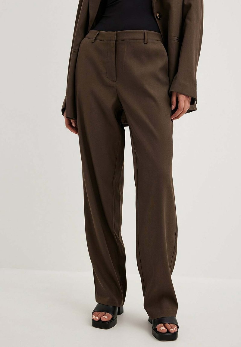 Brązowe, dopasowane spodnie z prostymi nogawkami, subtelnym połyskiem, kieszeniami z przodu i szlufkami na pasek, w zestawie z czarnymi sandałami na platformie.