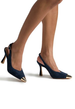 Talons hauts en suede bleu marine avec des embouts métalliques dorés pointus et une fine hauteur de talon. Équipés d'une boucle sur le côté pour un ajustement.