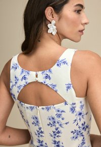 Blommig klänning i vit tyg med blå mönster, med en nyckelhålsknäppning och dragkedja. Stora blommiga örhängen ger detaljer.