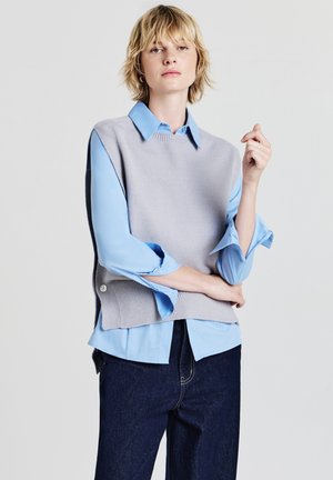 Gilet lavorato a maglia grigio chiaro sopra una camicia blu con colletto, caratterizzato da un orlo a coste e bottoni laterali, abbinato a pantaloni di denim scuri.