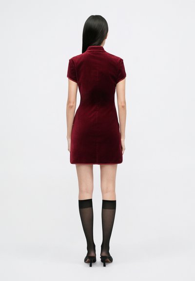 Rochie burgundă din catifea, cu mâneci scurte, guler și o siluetă ajustată, combinată cu șosete negre transparente până la genunchi și toc.