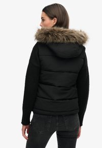 Frau, die seitlich steht, trägt eine schwarze Steppweste mit einer Kapuze aus Kunstpelz und schwarzen Strickärmeln, kombiniert mit dunkelgrauen Jeans.