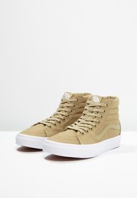 Ein Paar beige High-Top Vans Sneaker mit weißen Gummisohlen und passenden beigen Schnürsenkeln auf einer weißen Oberfläche mit schlichtem Hintergrund.