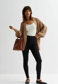 Cárdigan de punto marrón sobre una camiseta sin mangas blanca, combinado con jeans skinny negros y bailarina negra, sosteniendo un bolso marrón, sobre un fondo claro.