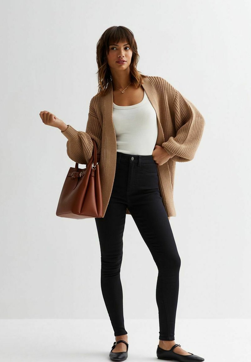 Cárdigan de punto marrón sobre una camiseta sin mangas blanca, combinado con jeans skinny negros y bailarina negra, sosteniendo un bolso marrón, sobre un fondo claro.