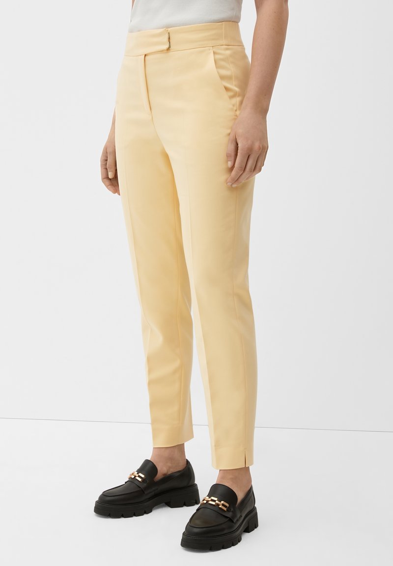 Pantalon sur mesure jaune clair avec une texture lisse, coupe fuselée et plis avant. Porté avec des mocassins noirs ornés d'accents de chaînes dorées.