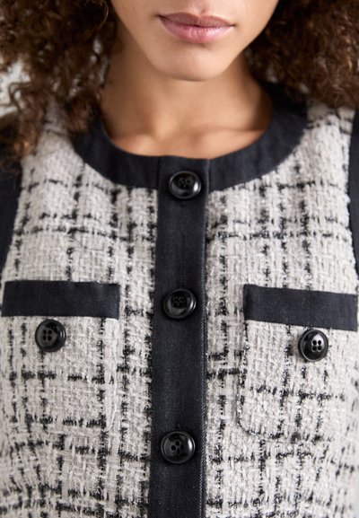 Veste en tweed présentant un mélange de tons gris et noirs, avec quatre boutons noirs et deux poches avant. Tissu texturé avec un design structuré.