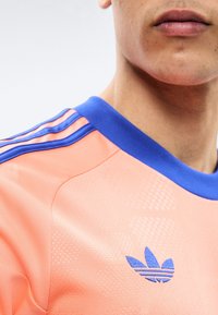 Približni prikaz osobe koja nosi sportsku majicu boje lososa s plavim ovratnikom i tri plave pruge na ramenu, prikazujući dio lica i vrata.