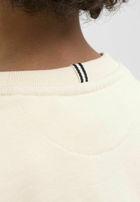 Krämfärgad sweatshirt med ribbat krage och en svart dubbelrand på baksidan, texturerad tyg och subtila stygn detaljer.