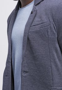 Grå stickad kavaj med slag, enkel knappstängning och bröstficka, bärs över en ljusblå strukturell rundhalsad tröja.