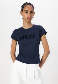Mėlynos spalvos trumpa rankovė T-shirt su tekstūriniu „GUESS“ logotipu tamsioje medžiagoje. Turi apvalų iškirpimą ir prigludusią siluetą.