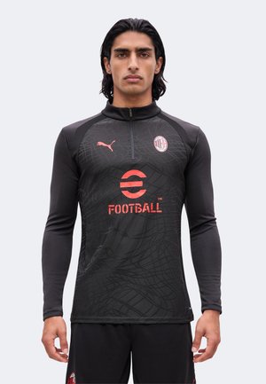 Mężczyzna o ciemnych włosach nosi czarną koszulkę piłkarską AC Milan z różowym logo Puma i czerwonym emblematem piłkarskim, stoi na tle jednolitego jasnego tła.