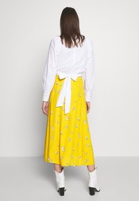 Chemise blanche à manches longues avec un détail noué à l'arrière, associée à une jupe florale jaune vif et des bottines blanches à talon bloc.