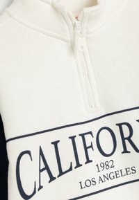 Sweatshirt crème avec un col montant et fermeture éclair. Présente un texte bleu marine "CALIFORNIA 1982 LOS ANGELES" et une texture douce.