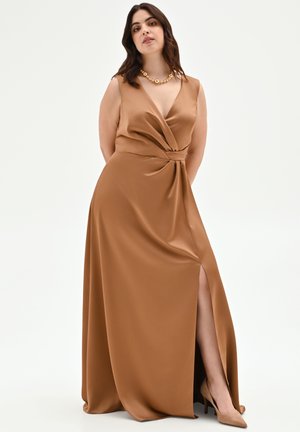 Donna in un abito marrone in raso senza maniche con scollatura a V profonda, spacco laterale, tacchi beige e una collana d'oro, in piedi con le mani dietro la schiena.