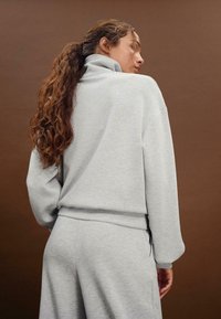 Jasnoszara, przytulna bluza z wysokim kołnierzem, z luźnymi rękawami i fakturowanym materiałem. Prosty design, połączony z pasującymi spodniami.