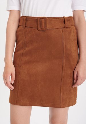 Bruine suède minirok met bijpassende riem en rechthoekige gesp, gedragen met een witte top, zichtbaar vanaf de taille tot halverwege de dij.