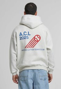 Hanorac cu glugă de culoare gri deschis, cu text grafic roșu și albastru și un motiv de tenis pe spate. Fabricat din bumbac moale, cu manșete ribbate.
