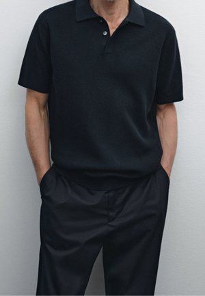 Man wearing a black short-sleeve textured polo shirt tucked into black high-waisted pants with hands in pockets, standing against plain background.