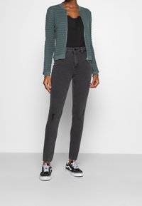 Turkooise doorgestikte bomberjack, zwart kanten tanktopje, versleten grijze spijkerbroek en zwart-witte sneakers. Strak en eigentijds ontwerp.