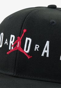 Svart caps med hevet rødt "Jumpman"-logo og hvit brodert tekst. Designet inkluderer tekstil med struktur og bøyd skygge.