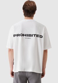 White cotton t-shirt featuring large black text "PROHIBITED" and "10119, BERLIN" on the back. Relaxed fit, short sleeves, straight hem.