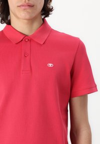 Jeune personne portant un polo à manches courtes de couleur rouge vif avec deux boutons et un petit logo brodé blanc sur la poitrine.