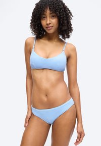 Γυναίκα με κατσαρά μαλλιά, που φοράει ένα υφασμάτινο ανοιχτό μπλε μπικίνι, στέκεται με το ένα χέρι χαλαρό στο πλάι, πάνω σε λευκό φόντο.