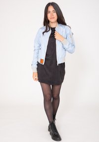 Ljusblå bomberjacka med grå mudd och en orange etikett. Bärs över en svart klänning och matchas med svarta snörboots. Slät textur.