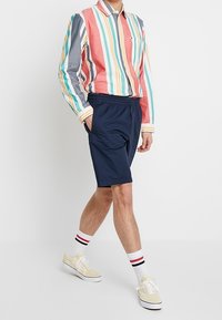 Pestrý pruhovaný bavlněný shirt, kombinovaný s námořnickými kraťasy. Outfit obsahuje bílé ponožky s červenými a černými pruhy a žluté tenisky.