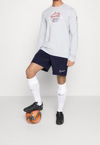 Camisa de manga larga gris claro con el logo rojo del Liverpool FC, pantalones cortos azul marino, calcetines blancos, botas de fútbol negras, de pie sobre un balón de fútbol naranja.